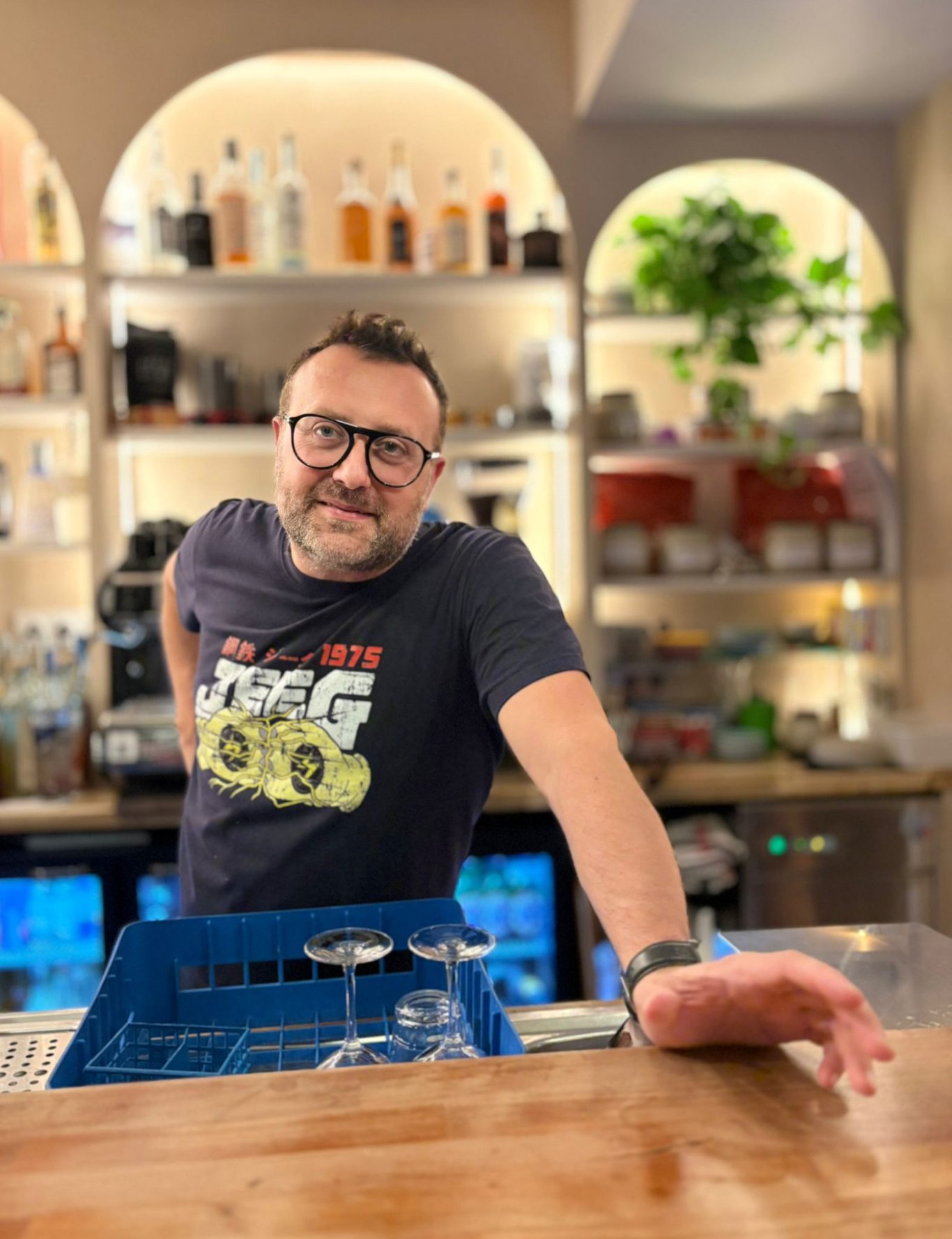 Homme souriant derrière un bar, avec des bouteilles et des verres en arrière-plan.