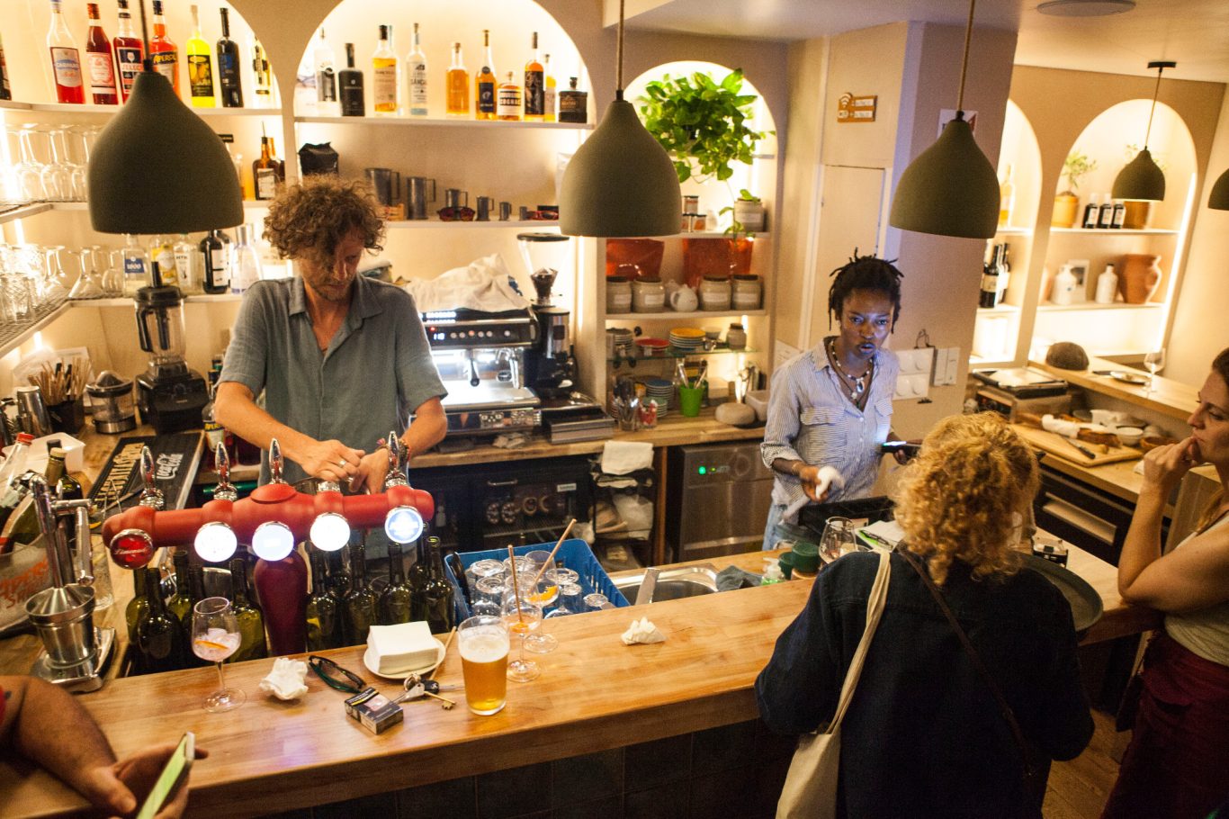 Deux barmans préparent des boissons derrière un bar animé, des clients en attente.