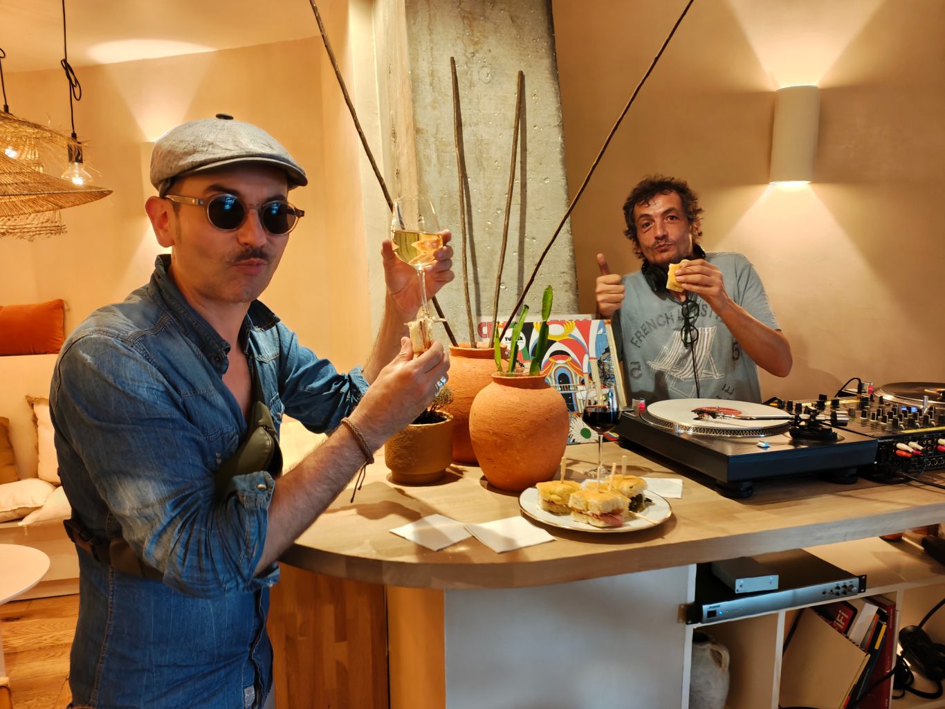 Deux hommes, l'un avec des lunettes de soleil, lèvent un verre près d'une platine de DJ.