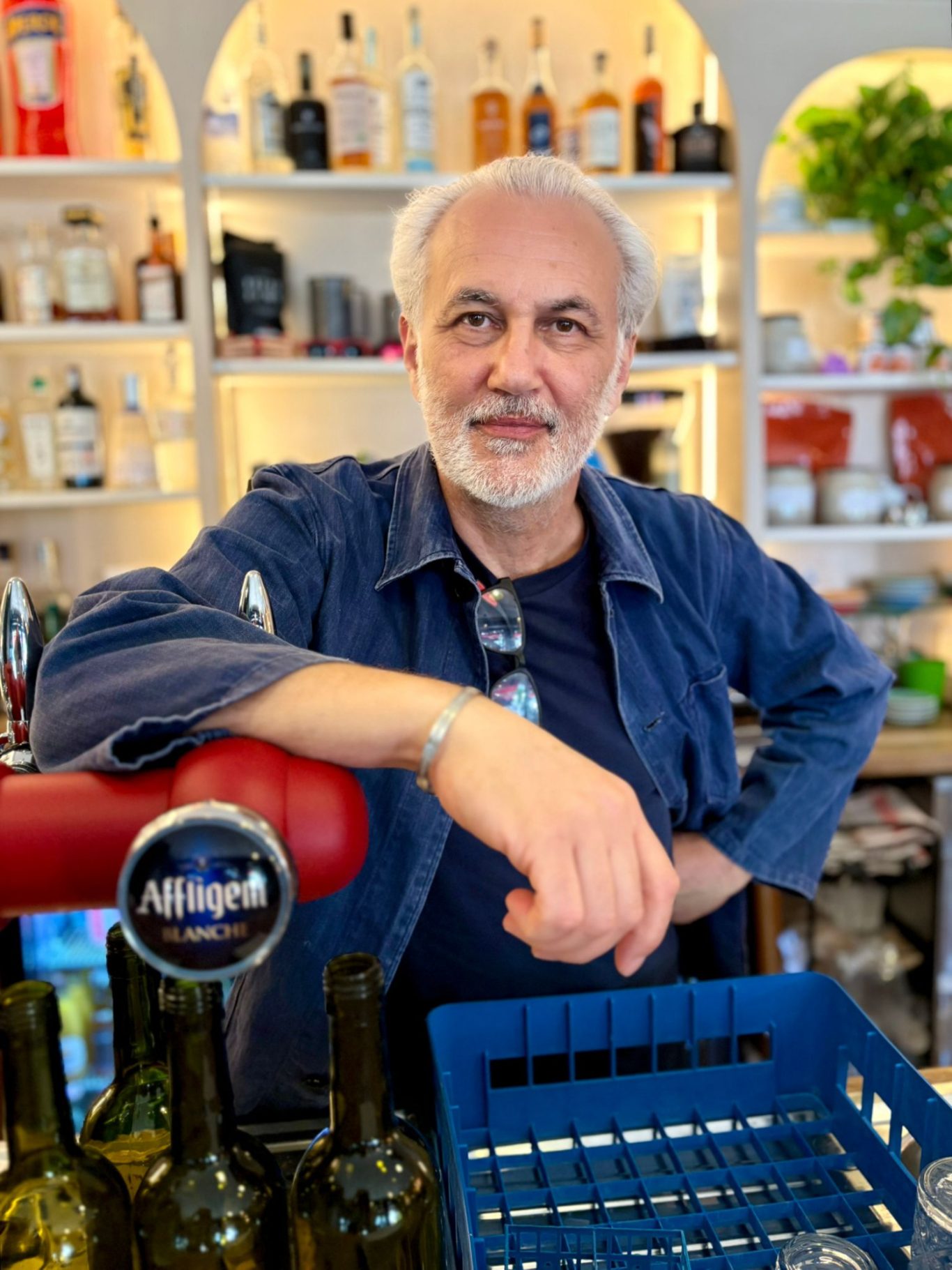 Homme souriant avec une chemise bleue, devant un magasin bien approvisionné.