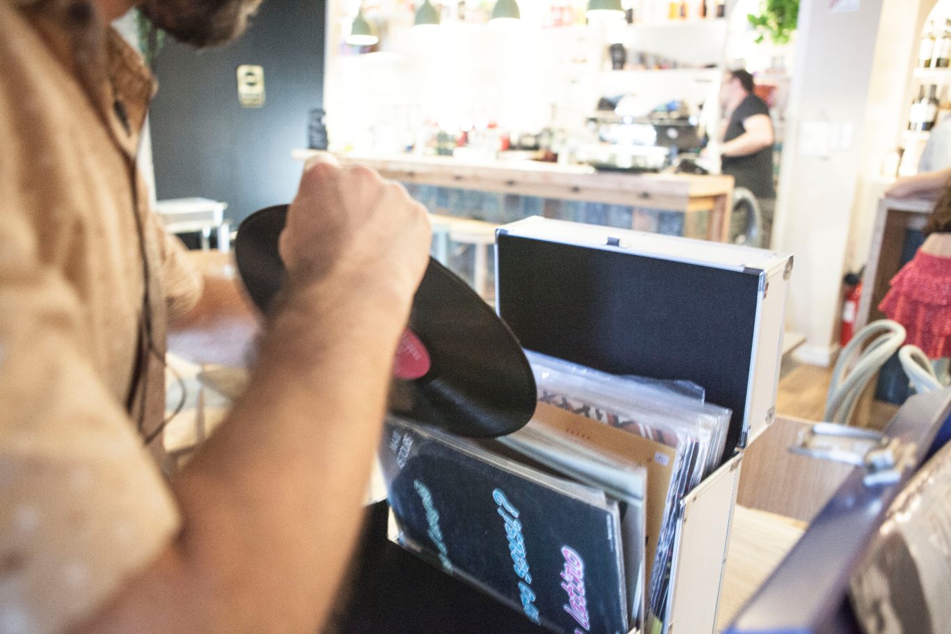 Homme feuilletant des disques vinyles dans une boîte, ambiance de café en arrière-plan.