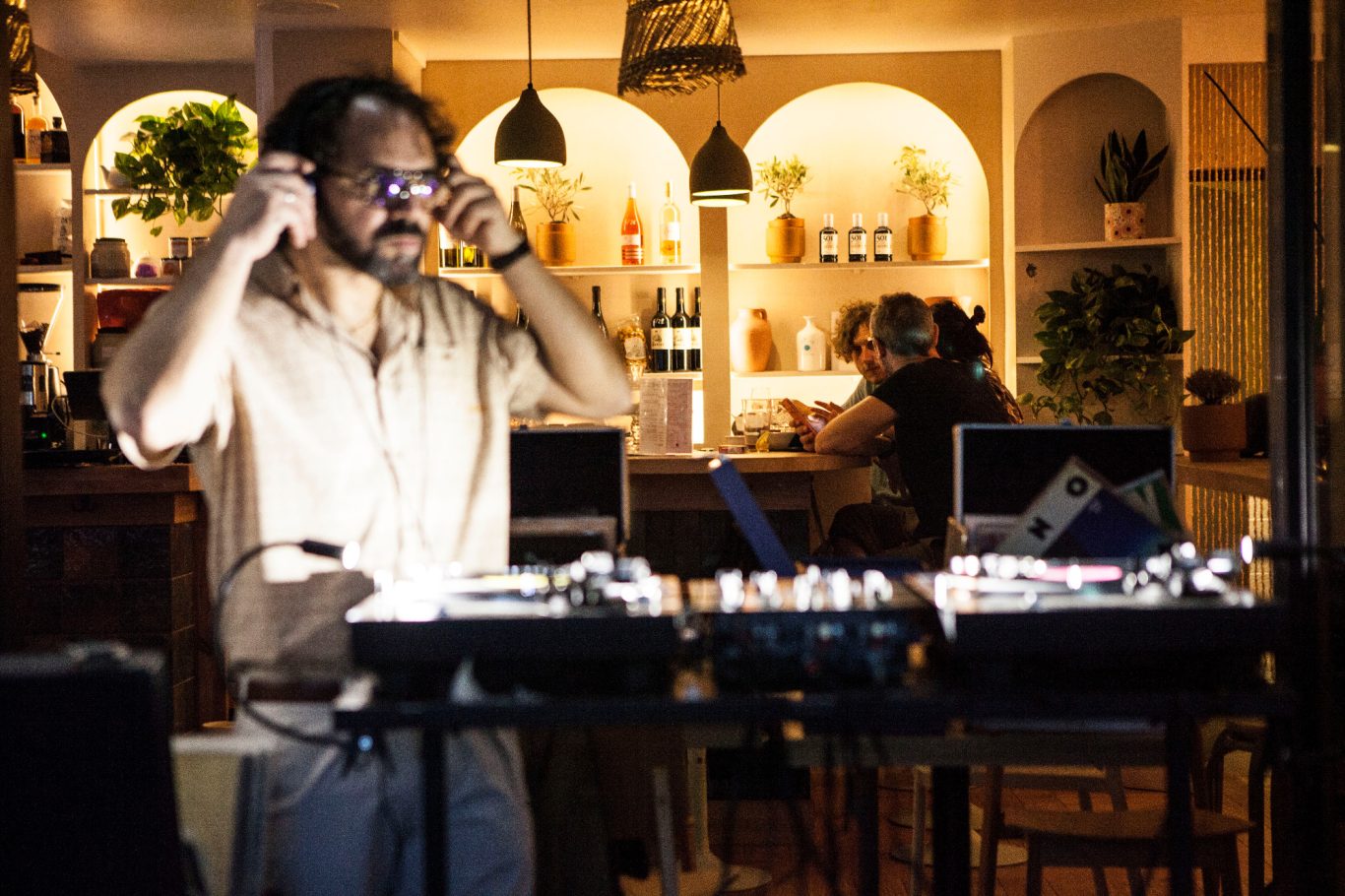 Un homme avec des lunettes ajuste son casque devant des platines dans un bar animé.