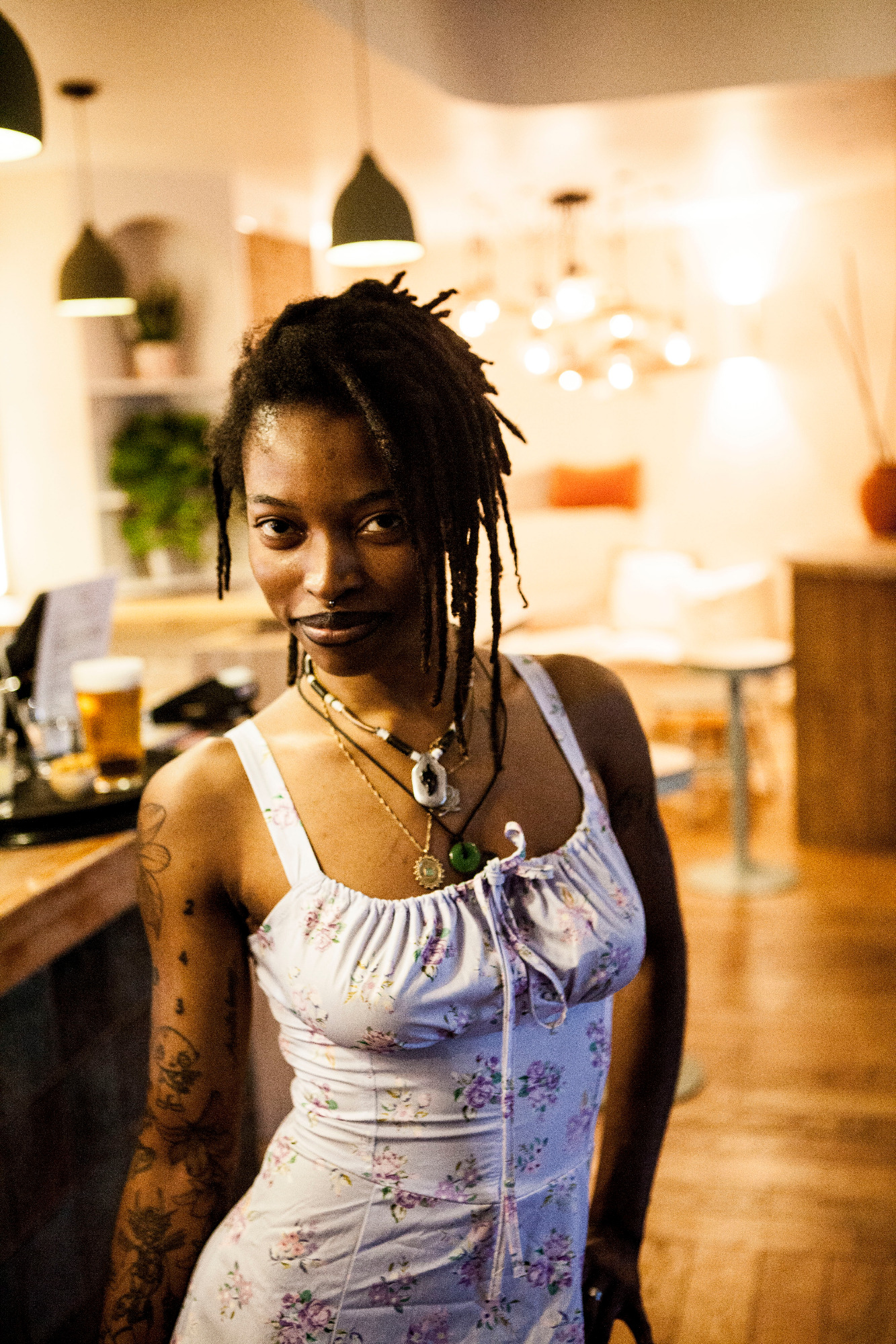 Femme à la peau foncée avec des dreadlocks, vêtue d'un haut léger, dans un café lumineux.